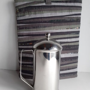May include: A silver stainless steel French press coffee maker with a lid and handle. The coffee maker is sitting on a gray and white striped fabric surface.
