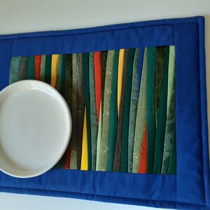 May include: A blue quilted placemat with a colorful striped design in the center. The placemat has a white plate on top.