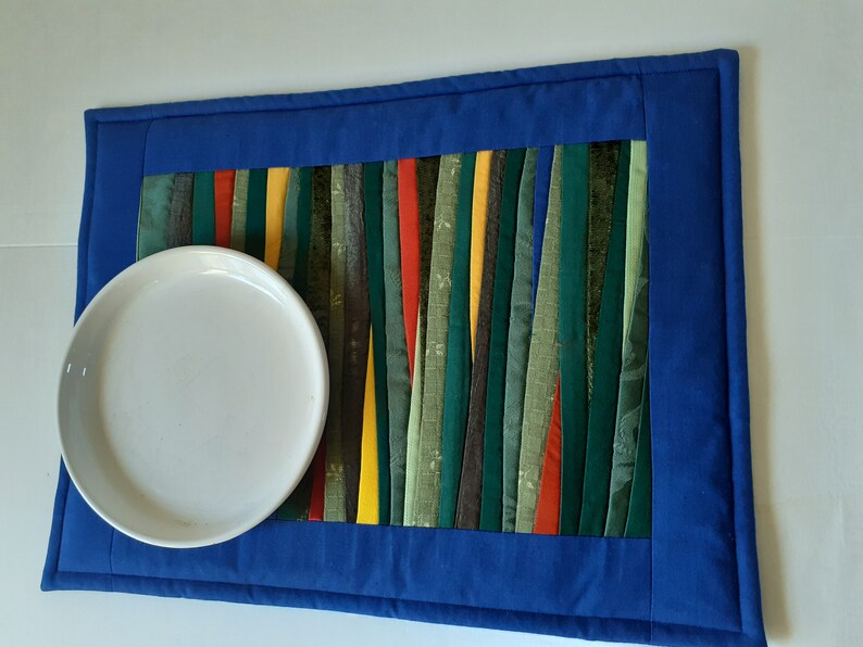 May include: A blue quilted placemat with a colorful striped design in the center. The placemat has a white plate on top.