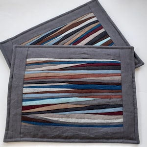 May include: Two rectangular placemats with a grey border and a patchwork design. The patchwork features horizontal stripes in shades of blue, brown, and grey. The placemats are made of fabric and are suitable for dining.