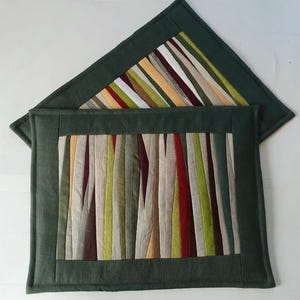 May include: Two handmade placemats with a forest green border. The center of each placemat features vertical stripes in various colors, including red, green, beige, and yellow. The top placemat is triangular, and the bottom one is rectangular.