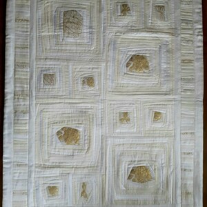 Könnte beinhalten: Cremefarbene Steppdecke mit geometrischem Design. Die Steppdecke zeigt Quadrate in Quadraten, mit goldfarbenen Formen in der Mitte einiger Quadrate. Der Rand ist eine hellere cremefarbene Farbe mit vertikalen Streifen.