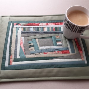 May include: A green quilted placemat with a geometric pattern of various shades of gray, blue, and pink fabric. A white mug with black stripes sits on the placemat.