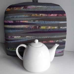 Peut inclure: Une théière blanche avec un couvercle est placée devant un chauffe-thé en tissu matelassé gris et violet avec une anse. Le chauffe-thé a un motif rayé avec des accents floraux.