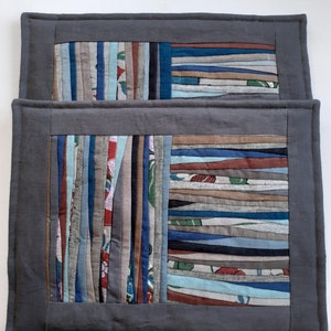 Puede incluir: Dos manteles individuales de tela gris con un diseño de patchwork de tiras de tela de colores. Las tiras están dispuestas verticalmente y cosidas juntas para crear una forma rectangular.
