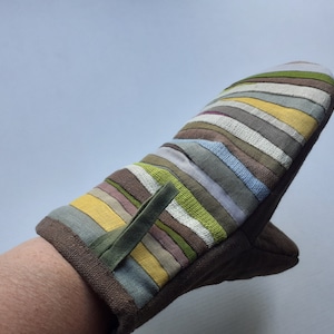 Pode incluir: Uma luva de forno acolchoada com exterior castanho e um padrão listrado em tons de verde, amarelo, cinza e roxo. Possui uma alça para pendurar e é projetada para uso na cozinha.