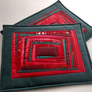 以下が含まれることがあります： ログキャビンデザインの正方形のキルトプレースマット2枚。中央には赤とピンクの生地のらせん模様があり、濃いティール色のボーダーで縁取られています。プレースマットは手作りです。