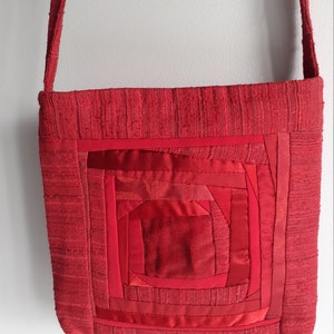 Może przedstawiać: Czerwona torba na ramię o kwadratowym kształcie i długim pasku. Torba posiada centralny wzór z koncentrycznych kwadratów w różnych odcieniach czerwieni, tworząc efekt patchworku. Torba wykonana jest z teksturowanej tkaniny.