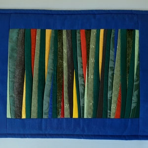 May include: A blue fabric placemat with a border and a center panel made of colorful fabric strips. The strips are arranged vertically and are in shades of green, yellow, red, and black.