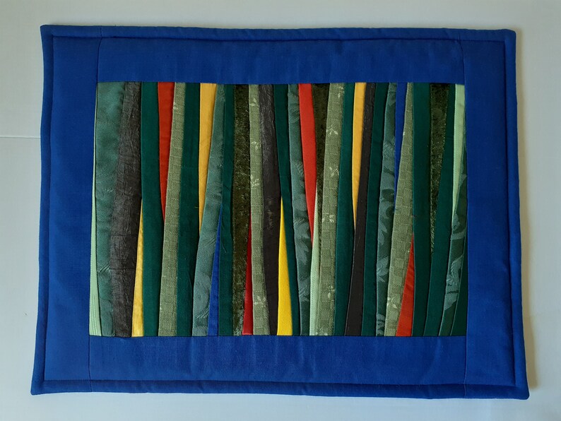 May include: A blue fabric placemat with a border and a center panel made of colorful fabric strips. The strips are arranged vertically and are in shades of green, yellow, red, and black.