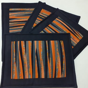 Op de afbeelding: Set van vijf zwarte placemats met oranje en grijze gestreepte stoffen inzetstukken. De stof is in dunne stroken geknipt en aan elkaar genaaid om een gestreept patroon te creëren.