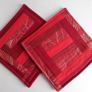 May include: Two square, quilted potholders in shades of red. The potholders feature a patchwork design with varying textures and patterns, including floral and linear elements. The edges are neatly finished with a darker red binding.
