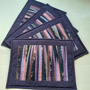 May include: A set of four rectangular quilted placemats featuring a central panel of vertical fabric strips in shades of purple, pink, brown, and black. Each placemat is framed by a solid dark purple border with visible machine stitching.