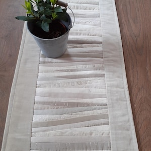 May include: Cream-colored table runner with a quilted design featuring horizontal strips of varying textures and shades of white and cream. A small metal bucket with a plant sits on the runner. The runner is approximately 12 inches wide.