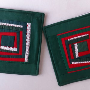Può includere: Due sottopentola quadrati in tessuto verde con un design a casetta di legno rosso e bianco. Il design presenta un bordo in pizzo bianco.