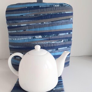 May include: A white teapot with a blue and grey striped tea cosy and matching coaster. The cosy is made of fabric strips sewn together.