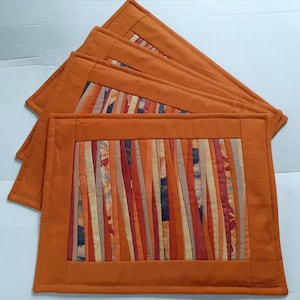 May include: A set of four rectangular placemats featuring a solid orange border surrounding a central panel of vertical fabric strips in shades of orange, red, yellow, and dark blue. The strips vary in width and are arranged in a linear pattern.