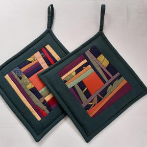 Może przedstawiać: Dwa kwadratowe, pikowane podkładki pod garnki z ciemnozieloną obwódką i pętelką do zawieszania. Środek zdobi wzór patchworkowy z paskami w różnych kolorach, w tym pomarańczowym, fioletowym i zielonym. Do użytku w kuchni.