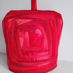 May include: Red tea cosy with a quilted rose design made from various shades of red fabric.