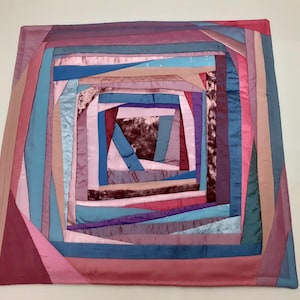 May include: A square pillow with a geometric pattern made of pink, blue, and purple fabric. The fabric is arranged in a concentric square pattern, with the center of the pillow being the smallest square.