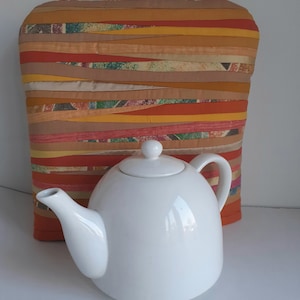 Puede incluir: Una tetera de cerámica blanca con tapa y asa, sentada sobre un calienta-tetera de tela a rayas de colores. El calienta-tetera está hecho de tiras de tela de varios tonos de naranja, marrón y beige.