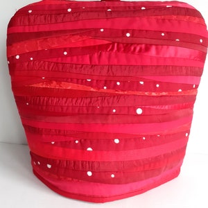 May include: Red and pink teapot cosy with white polka dots. The cosy is made of fabric strips sewn together in a wavy pattern.