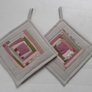 Op de afbeelding: Twee vierkante pannenlappen met een lichtgrijze rand en een centraal patchworkontwerp. Het patchwork heeft roze, groene en bloemenstoffen in een geometrisch patroon. Elke pannenlap heeft een kleine stoffen lus om op te hangen.