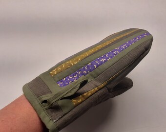 Puede incluir: Un guante de horno verde con rayas moradas y doradas. El guante tiene un lazo en la muñeca para colgarlo.