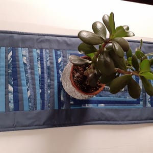 May include: A blue quilted table runner with a patchwork design of various shades of blue. A small potted plant with dark green leaves sits on the runner. The runner is rectangular and has a solid blue border.