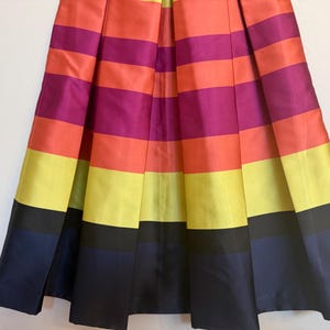 May include: A pleated skirt with horizontal stripes in vibrant colors: orange, fuchsia, yellow, and navy blue. The skirt has a satin-like sheen, creating a bold and eye-catching design. The skirt is a knee-length style.