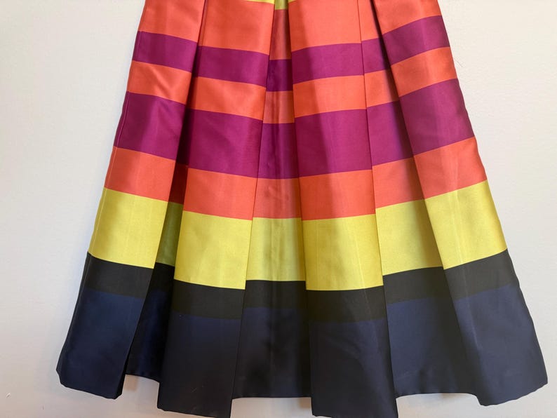 May include: A pleated skirt with horizontal stripes in vibrant colors: orange, fuchsia, yellow, and navy blue. The skirt has a satin-like sheen, creating a bold and eye-catching design. The skirt is a knee-length style.