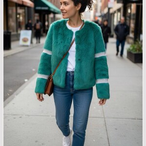 Op de afbeelding: Een vrouw draagt een teal imitatiebontjas met witte strepen op de mouwen en zoom, gecombineerd met een blauwe jeans en witte sneakers. Ze draagt een bruine tas en loopt op een stoep.