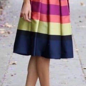 May include: A knee-length, pleated skirt with horizontal stripes in shades of red, purple, yellow, and navy blue. The skirt is paired with black high heels. The outfit is completed with a white shirt and a black belt.