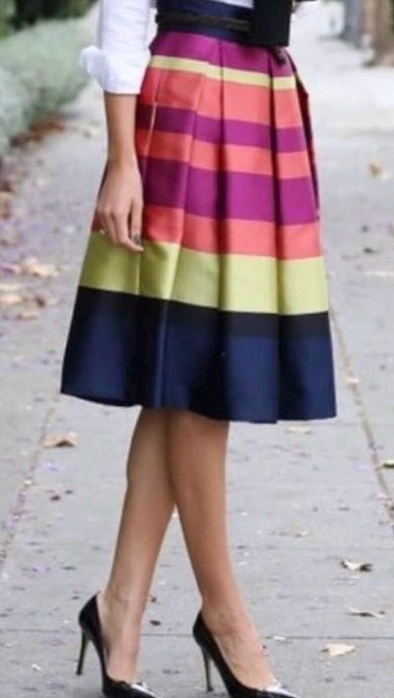 May include: A knee-length, pleated skirt with horizontal stripes in shades of red, purple, yellow, and navy blue. The skirt is paired with black high heels. The outfit is completed with a white shirt and a black belt.
