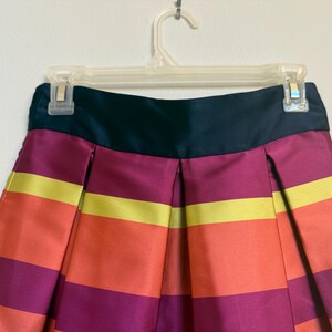 May include: A colorful skirt with horizontal stripes in shades of orange, yellow, and purple. The skirt has a dark teal waistband and is hanging on a clear plastic hanger. The fabric appears to be satin or a similar material.