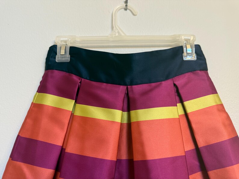 May include: A colorful skirt with horizontal stripes in shades of orange, yellow, and purple. The skirt has a dark teal waistband and is hanging on a clear plastic hanger. The fabric appears to be satin or a similar material.