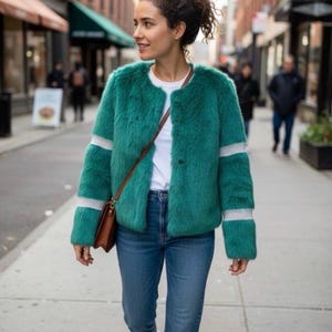 Op de afbeelding: Een vrouw draagt een teal imitatiebontjas met witte strepen op de mouwen, een wit t-shirt en een blauwe jeans. Ze draagt een bruine leren crossbodytas en witte sneakers. De achtergrond toont een stadsstraat.