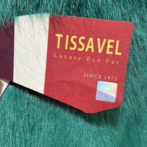 Op de afbeelding: Close-up van een label op een teal imitatiebont stof. Het label is rood met de woorden "TISSA VEL Luxury Eco Fur" in gouden letters. Het label zegt ook "Since 1975" en "Originated in France".