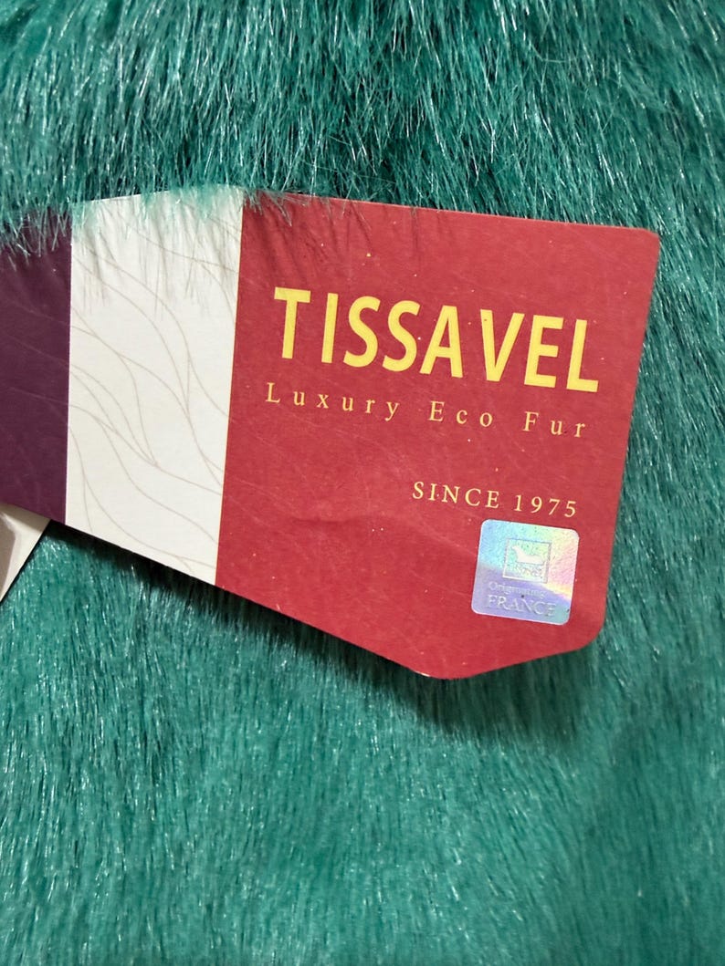 Op de afbeelding: Close-up van een label op een teal imitatiebont stof. Het label is rood met de woorden "TISSA VEL Luxury Eco Fur" in gouden letters. Het label zegt ook "Since 1975" en "Originated in France".