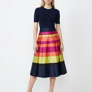 May include: A navy blue short-sleeve top paired with a vibrant, pleated midi skirt. The skirt features horizontal stripes in shades of pink, orange, yellow, and navy blue. The model is wearing nude-colored heels.