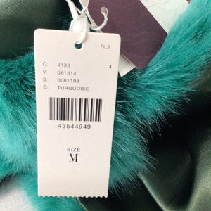 Op de afbeelding: Een close-up van een label met de tekst "TURQUOISE" en "SIZE M". Het label is bevestigd aan een turquoise imitatiebontafwerking en een donkergroene satijnen stof. De barcode is zichtbaar.