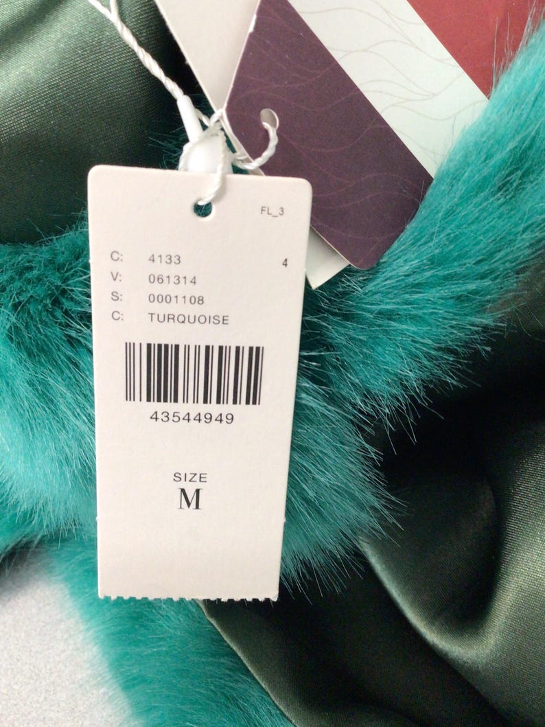 Op de afbeelding: Een close-up van een label met de tekst "TURQUOISE" en "SIZE M". Het label is bevestigd aan een turquoise imitatiebontafwerking en een donkergroene satijnen stof. De barcode is zichtbaar.