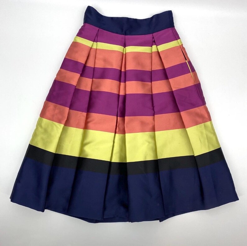May include: A pleated skirt with horizontal stripes in shades of navy, black, yellow, coral, purple, and pink. The skirt has a navy waistband and a full, A-line silhouette. The fabric appears to have a slight sheen.