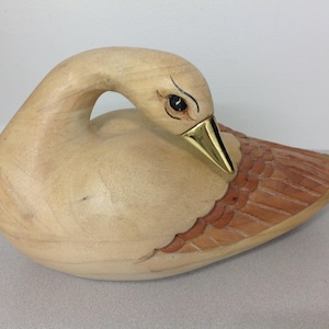 May include: Wooden swan sculpture with a curved neck and detailed wing design. The swan has a light wood body, a brass beak, and brown carved wings. The eye is painted black. A decorative piece for home decor.