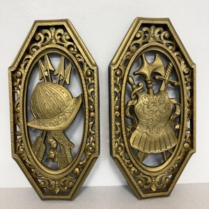 May include: A pair of gold-colored wall plaques with an octagonal frame and oval center. The left plaque features a helmet and arrows, while the right plaque displays a breastplate, a sword, and an axe, all rendered in a relief design.