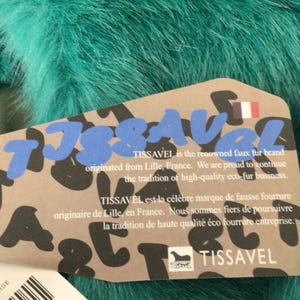 Op de afbeelding: Close-up van teal imitatiebont met een productlabel. Het label toont de merknaam "TISSAVEL" in blauwe en zwarte letters, met tekst in het Engels en Frans. Het merk komt uit Lille, Frankrijk.