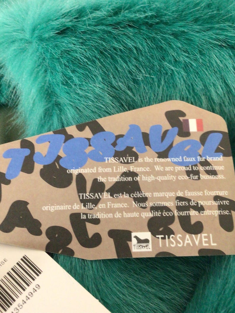 Op de afbeelding: Close-up van teal imitatiebont met een productlabel. Het label toont de merknaam "TISSAVEL" in blauwe en zwarte letters, met tekst in het Engels en Frans. Het merk komt uit Lille, Frankrijk.