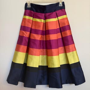 May include: A pleated skirt with horizontal stripes in shades of purple, orange, yellow, and navy blue. The skirt is hanging on a white hanger against a white background. The skirt has a navy blue waistband.