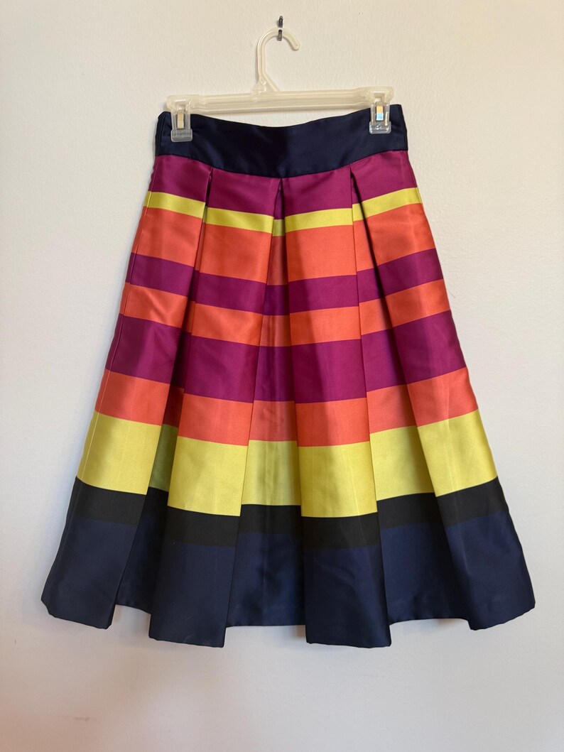 May include: A pleated skirt with horizontal stripes in shades of purple, orange, yellow, and navy blue. The skirt is hanging on a white hanger against a white background. The skirt has a navy blue waistband.