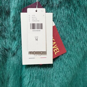 Op de afbeelding: Close-up van turquoise imitatiebontstof. Een wit label met zwarte tekst en een barcode is bevestigd, met de letter "M" voor de maat. Een rood label met het woord "CAVEL" is ook zichtbaar.